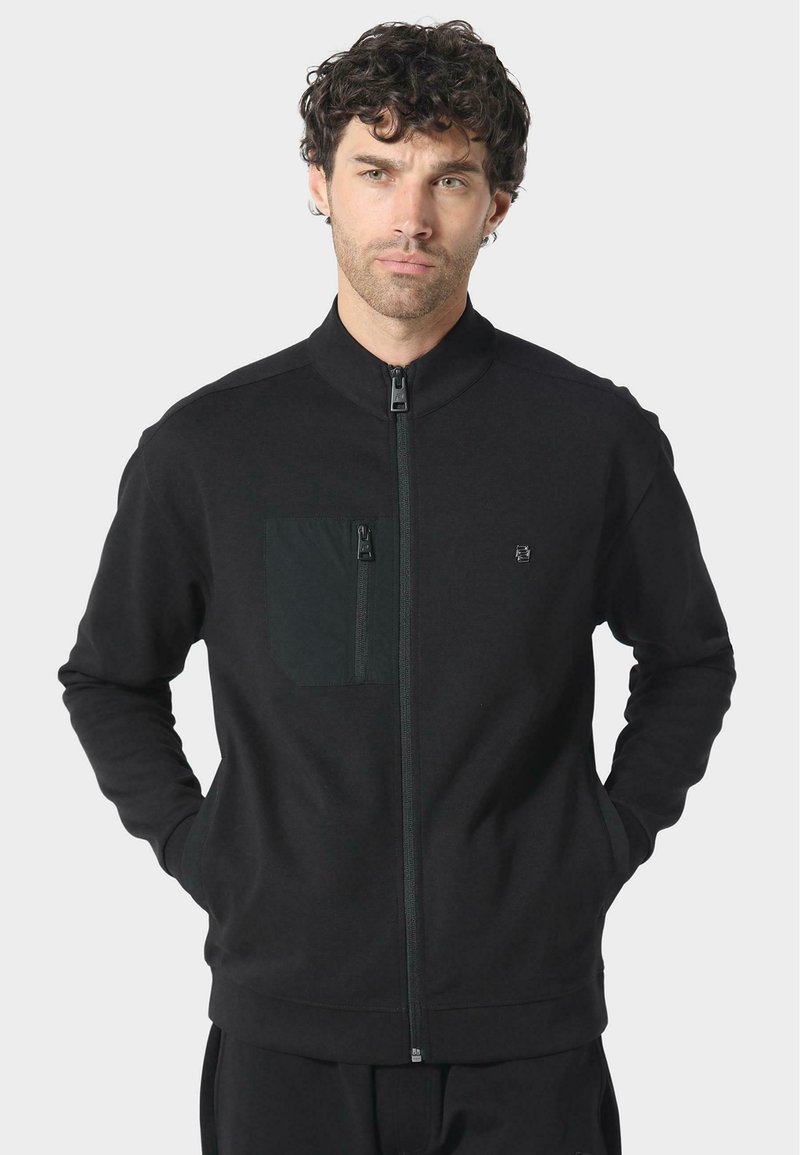 Černá zipová bunda vyrobená z měkké tkaniny, s kapsou na zip na hrudi, vysokým límcem a žebrovanými manžetami. Jednoduchý design, bez vzorů.