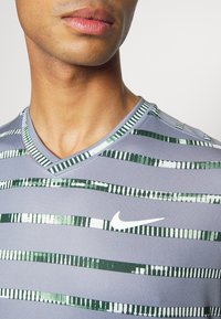 Camiseta de cuello en V de Nike en gris con franjas horizontales verdes. El material es suave y tiene un corte ajustado. Logo de Nike en blanco en el pecho.