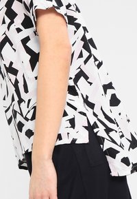 Camicia a maniche corte in un tessuto leggero che presenta un motivo geometrico astratto in bianco e nero con delicati accenti rosa e spacchi ai lati.