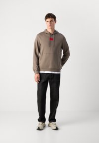 Felpa marrone con un patch logo rosso sul petto, tasca a marsupio frontale, abbinata a jeans neri e scarpe chiare. Design e vestibilità semplici.