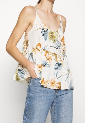 Kvinde iført en løs hvid blomster-ærmeløs top med tynde stropper og lyseblå denimjeans, hånd i lommen, foran en ensfarvet baggrund.