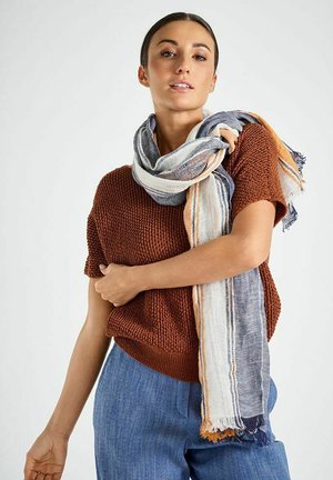 Femme portant un haut en maille couleur rouille et un pantalon bleu, ajustant une écharpe rayée multicolore drapée autour de son cou sur un fond clair.