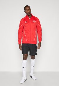 Red zip-up jacket with white accents and logo, black shorts, white socks with black logo, and white football boots. Smooth fabric texture.
