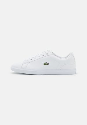 Zapatilla blanca hecha de material sintético con una textura suave, que presenta un logo bordado en verde en el lateral y un sutil diseño acolchado.