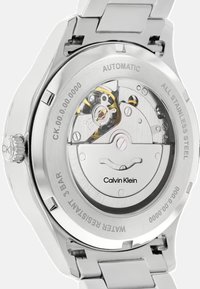 Automaatne roostevabast terasest kell hõbedase rihmaga. Märkimisväärne läbipaistev tagakaas, mis paljastab keerulised sisemehhanismid. Märgistatud "Calvin Klein".