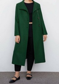 Personne portant un long manteau vert foncé croisé, un top court noir, un pantalon noir et des chaussures plates noires à clous.