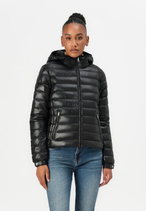 Svart pufferjakke med en blank finish, glidelås-lukking og hette. Har quiltet design og sidelommer. Kombinert med blå jeans.
