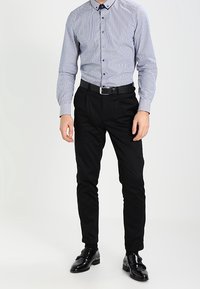 Pruhovaná modro-bílá košile na knoflíky s límečkem, doplněná úzkými černými kalhotami a lesklými černými botami s černým opaskem.