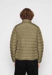 Olivgrön dunjacka med horisontellt quiltmönster, upprätt krage och strukturerad yta, kombinerad med svarta manchesterbyxor.