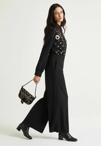 Pantaloni neri a gamba larga abbinati a una maglia nera decorata. Accessori comprendono una piccola borsa nera con motivi floreali e stivali con tacco.