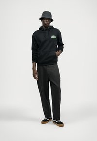 Jauns vīrietis, ģērbies melnā kausa cepurē, melnā kapučjakā ar zaļu logo, brīvās tumšās džinsās un melnās kedās ar baltām detaļām, stāv pret vienkāršu fonu.