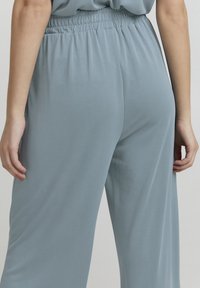 Primo piano di una persona che indossa pantaloni larghi con vita elastica di colore azzurro chiaro, con le mani rilassate ai lati, davanti a uno sfondo neutro.