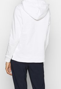 Sudadera blanca con capucha de diseño limpio, que cuenta con un bolsillo tipo canguro y puños acanalados, combinada con pantalones oscuros.