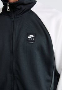 Crno-bijela Nike Air zip jakna s visokim ovratnikom, glatkom teksturom tkanine i kvadratnom logom oznakom na prsima.