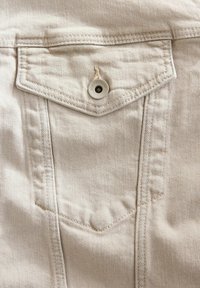Beige denim stof met een diamantvormige borstzak. Voorzien van een ronde, witte knop met de tekst "DENIM SUPPLY CO" en zichtbare stiksels.