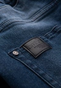 Jeans in denim blu scuro con una patch in pelle nera visibile con branding impresso e un dettaglio in metallo argentato sul bottone.