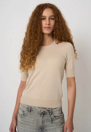 Femme aux longs cheveux roux bouclés portant un pull beige à manches courtes et un jean gris clair avec une ceinture à double boucle, debout devant un fond uni.