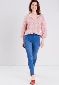 Rote Bluse mit floralem Muster, V-Ausschnitt, Puffärmeln und gerafftem Saum, kombiniert mit blauen Skinny Jeans und roten Sandalen.
