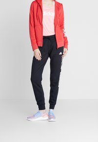 Sweat à capuche rouge zippé, t-shirt rose avec le logo "Adidas", jogging noir avec des rayures sur les côtés, et chaussures bleues avec des accents colorés.