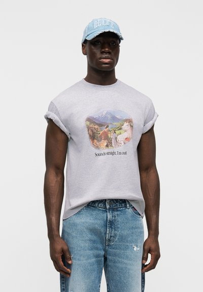 Sivá nadrozmerná tričko s rolovanými rukávmi a grafickou potlačou; kombinované s svetlomodrými roztrhanými džínsami a džínsovou čiapkou.