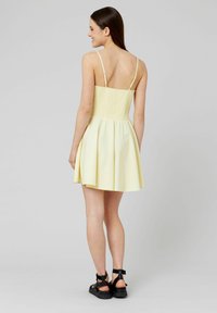 Robe d'été jaune avec un corsage froncé et une jupe évasée, dotée de fines bretelles réglables et d'un design sans dos. Portée avec des sandales noires.
