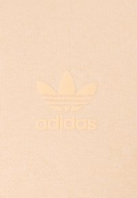 Beige bomullstyg med en upphöjd, tonad Adidas trefoil-logotyp med tre ränder under. Slät textur och minimalistisk design.