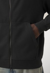 Sort zip-up hoodie med en blød tekstur, der har to sidelommer og ribbede manchetter, båret over lyse denim, designet til komfort.