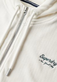 Crèmekleurige zip-up hoodie met geribbelde textuur, voorzien van een zilveren ritssluiting en een groen geborduurd "Superdry" logo op de borst.