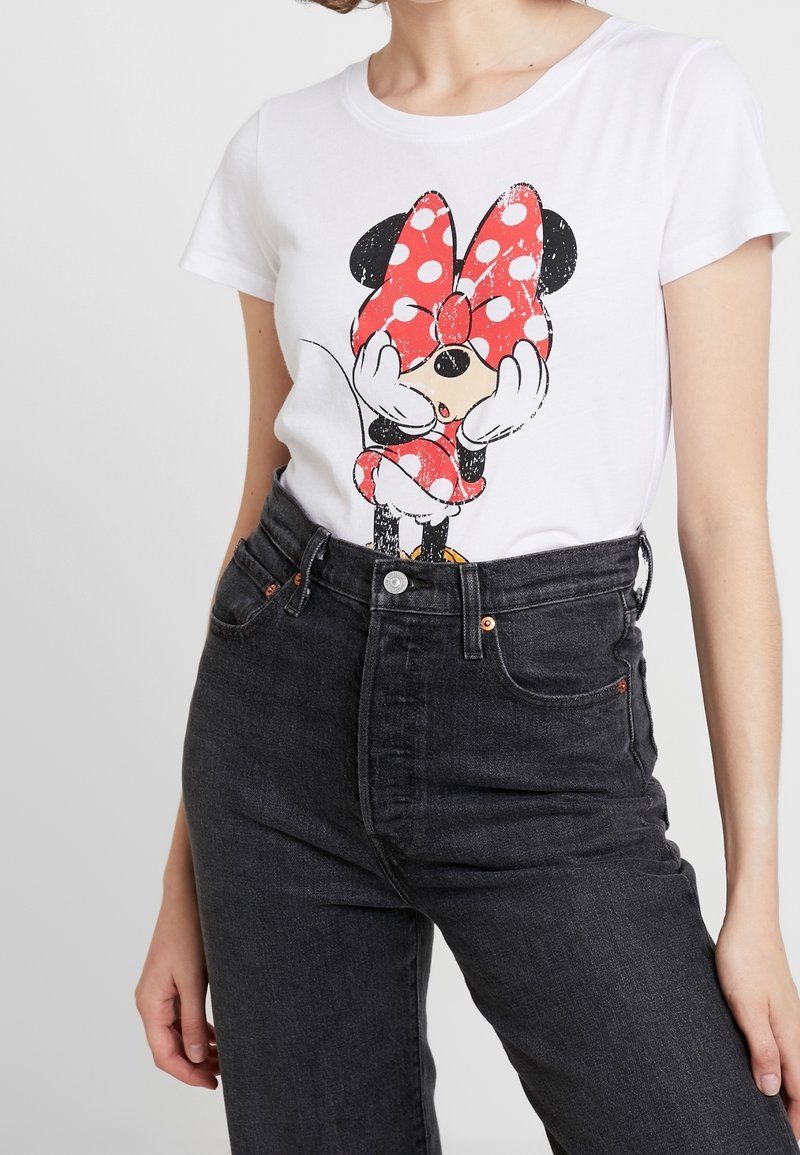 Femme portant un t-shirt blanc avec un motif Minnie Mouse couvrant ses yeux avec un grand nœud rouge à pois, associé à un jean noir taille haute.