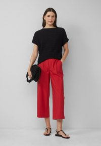 Haut noir texturé à manches courtes, pantalon rouge cropped, sac en bandoulière noir tressé et sandales plates noires. Tenue simple et décontractée avec des couleurs contrastées.