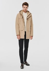 Beżowa parka z kapturem, dwiema przednimi kieszeniami z klapami i czarnymi spodniami. Model ma na sobie kremowy sweter i ciemnobrązowe buty. Gładka tekstura.