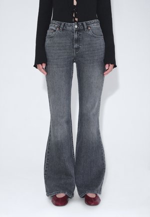 Osoba ubrana w wysokie, wyblakłe czarne dzwony, czarną bluzkę z długim rękawem z wiązaniami z przodu oraz ciemnoczerwone buty z kwadratowym noskiem.