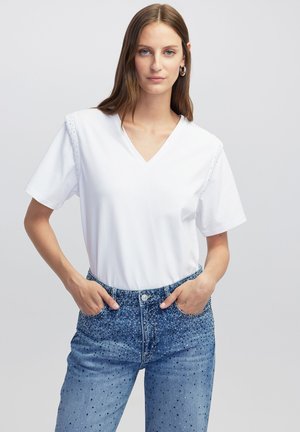 Witte katoenen t-shirt met een V-hals en korte mouwen, met gestikte accenten op de schouders. Gecombineerd met blauwe versierde denim jeans.