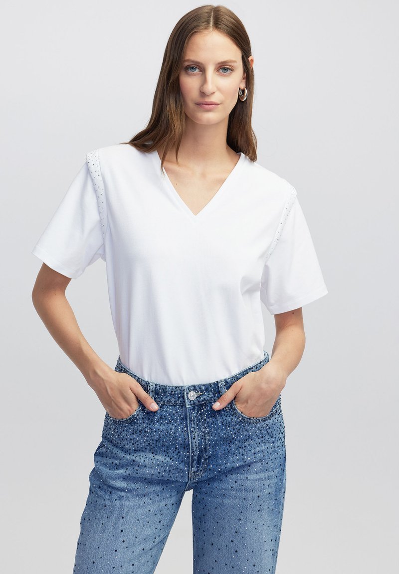 Witte katoenen t-shirt met een V-hals en korte mouwen, met gestikte accenten op de schouders. Gecombineerd met blauwe versierde denim jeans.
