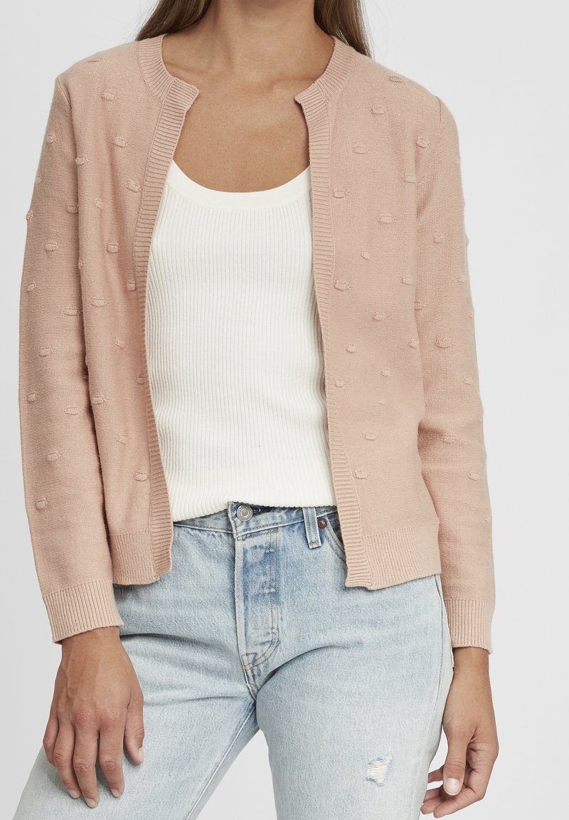 Cardigan lavorato a maglia di un rosa chiaro con design a pallini testurizzati, con apertura frontale, colletto e polsini a coste, indossato sopra una canotta bianca a coste.
