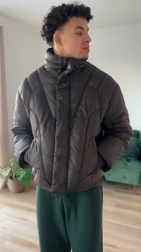 Donzen jas in donkerbruin, met een hoge kraag, doorgestikte textuur en ritssluiting. Twee zijzakken zijn zichtbaar.