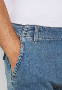 Jeans de mezclilla azul claro con costuras naranjas, que cuentan con un bolsillo delantero y una cinturilla clásica. La textura es suave con un ajuste casual.