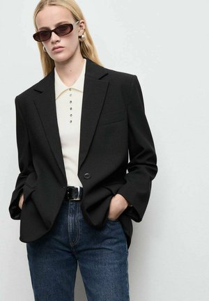 Blazer noir sur mesure avec un seul bouton, associé à un polo de couleur claire avec des détails de boutons et un jean bleu. Les accessoires incluent des lunettes de soleil sombres.