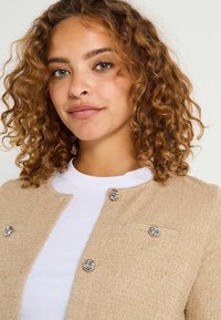 Cárdigan beige texturizado con cuello redondo, seis botones plateados en espiral y un detalle de bolsillo en el lado izquierdo, llevado sobre una camisa blanca.