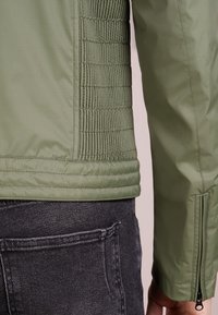 Veste vert clair avec des accents côtelés texturés sur le côté, tissu lisse sur les manches et une poche zippée au niveau du poignet.