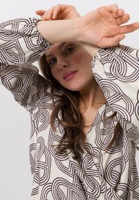 Witte blouse met een bruin abstract patroon, voorzien van elastische manchetten en een V-hals. De stof lijkt zacht en glad.