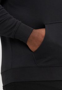 Fekete kapucnis pulóver hosszú ujjal, sima anyagból készült. Kanguruzsebbel és bordás mandzsettákkal rendelkezik, a kéz és az ujja közeli nézetét mutatja.