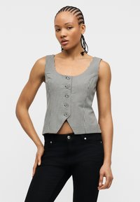 Γυναίκα με πλεξούδες, φοράει αμάνικο πουκάμισο σε μαύρο και λευκό καρό με κουμπιά και μαύρο παντελόνι, στέκεται μπροστά σε απλό φόντο.