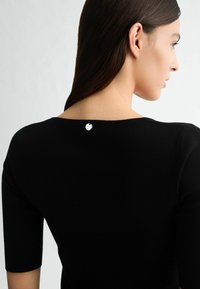 Schwarzes Strickoberteil mit weitem Ausschnitt und Dreiviertelärmeln, mit einem kleinen silbernen Logo-Button hinten in der Nähe des Ausschnitts.