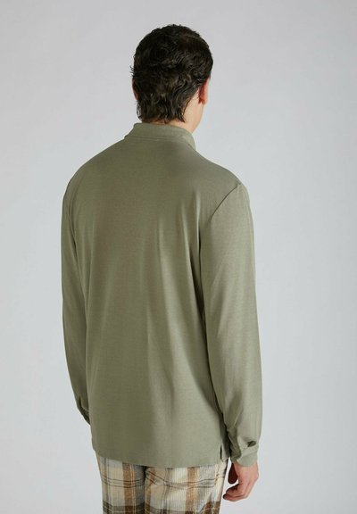 Camicia a maniche lunghe, di colore verde oliva, con colletto abbottonato. Presenta una texture liscia e fessure laterali. Indossata con pantaloni beige e marroni a fantasia.