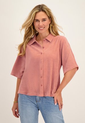 Vrouw met lang blond haar, gekleed in een doorgestikte stoffige roze blouse, lichtblauwe jeans, gouden oorringen en een armband, glimlachend naar de camera.