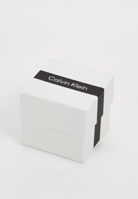 Weißer Geschenkkarton mit einem schwarzen Band, das das Etikett "Calvin Klein" trägt. Quadratische Form, glatte Oberfläche, minimalistisches Design.