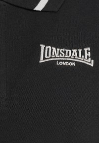 Camisa polo negra con tela texturizada, que presenta un cuello de rayas blancas y el logo bordado "Lonsdale London" en gris.