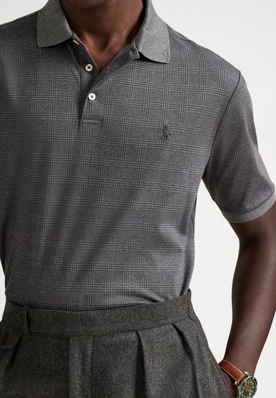 Άτομο με σκούρο δέρμα που φοράει γκρι καρό πόλο πουκάμισο κρυμμένο μέσα σε γκρι πλισέ παντελόνι, με καφέ ρολόι με δερμάτινο λουράκι στον καρπό.