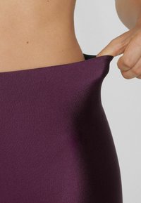 Leggings viola a vita alta con una texture liscia e elasticizzata, dotati di un ampio elastico che viene tirato leggermente verso il basso.